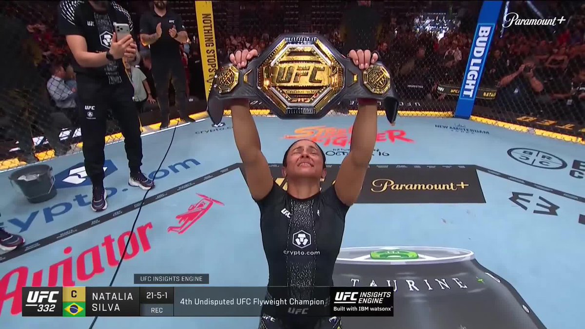 Brazilian fighter Natalia Silva (21-5-1) triumphantly hoists the UFC championship belt overhead inside the octagon after becoming the 4th Undisputed UFC Flyweight Champion at UFC 332, with broadcast graphics displaying her name, record, and the "UFC Insights Engine" banner as she celebrates her career-defining victory.