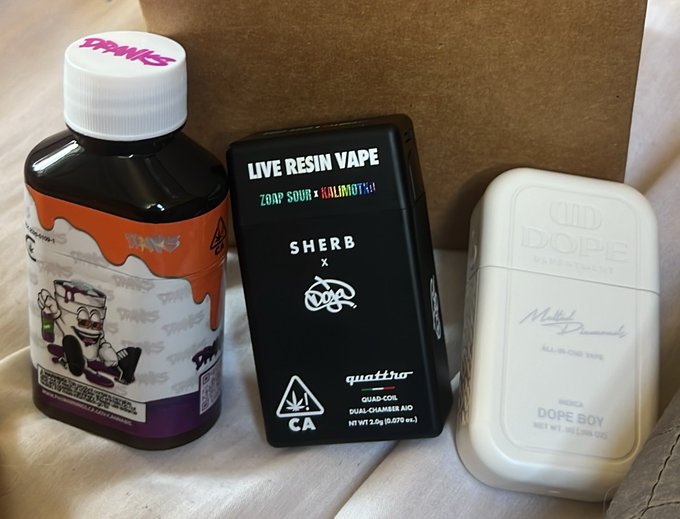 The image shows cannabis product packaging including a dark bottle labeled 'TRIPS', a black 'LIVE RESIN VAPE' box from 'ZAP SOUR x KALIMOTO' with 'SHERB x GEA' branding, and a white 'DOPE' 'DOPE BOY' container, all sitting inside a cardboard box.