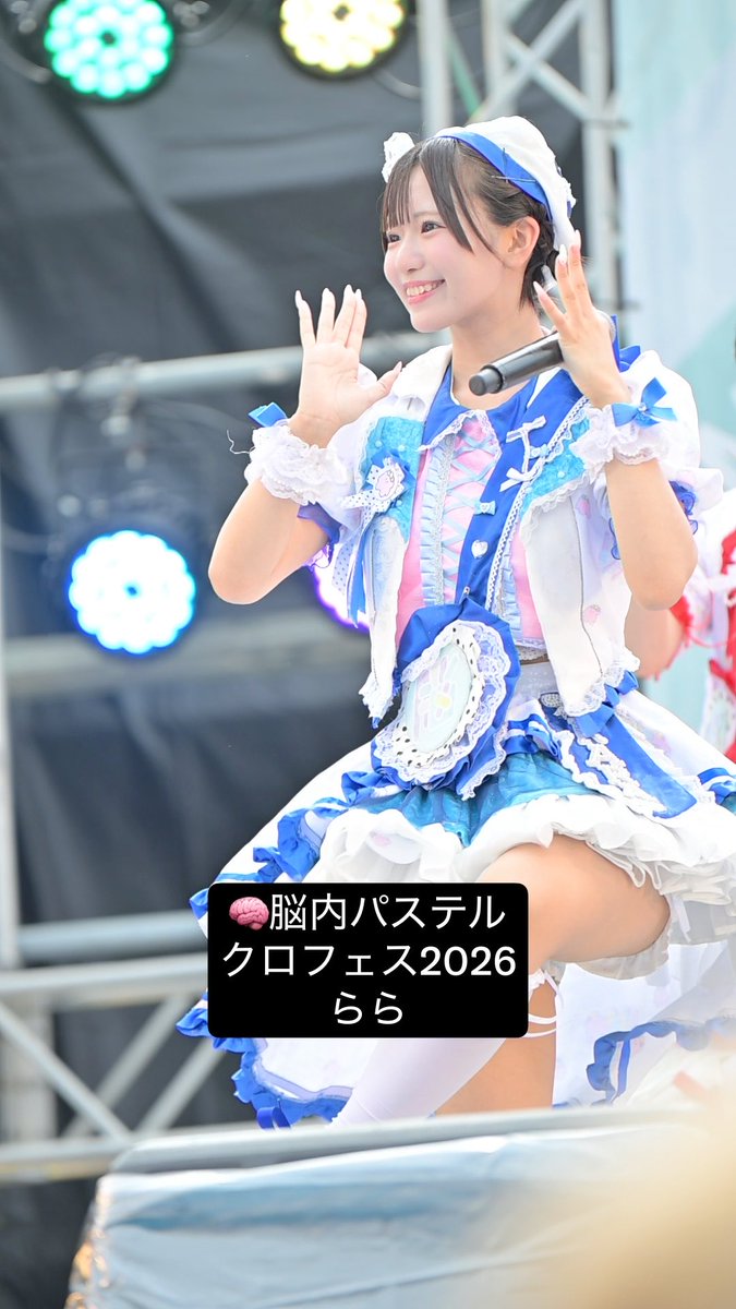 A performer on stage in a blue and white frilly idol costume, smiling and waving, with text overlay reading '脳内パステル クロフェス2026 らら'.