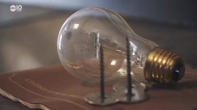 A vintage incandescent light bulb rests on a book, illustrating a product from before the Phoebus Cartel's 1925 decision to artificially limit bulb lifespans to 1,000 hours. The "abc 10" news logo is visible in the corner, suggesting this is from a news segment on planned obsolescence.