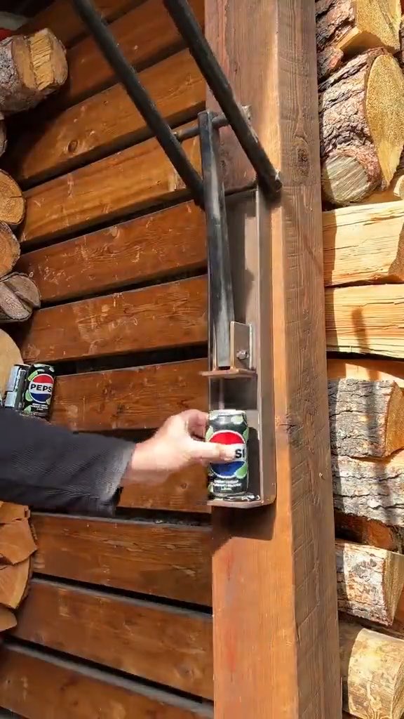 A person's hand places a can of Pepsi into a metal can holder mounted on a wooden wall next to a stack of firewood.
