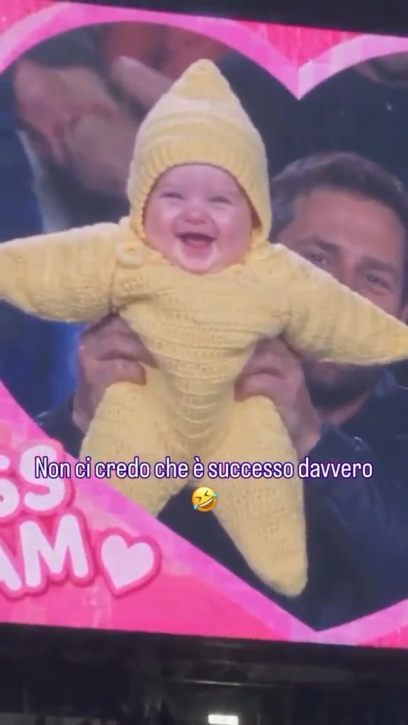 A baby wearing a yellow knitted star costume being held up in a crowd, smiling broadly, with Italian text overlay reading 'Non ci credo che è successo davvero' (I can't believe it really happened).