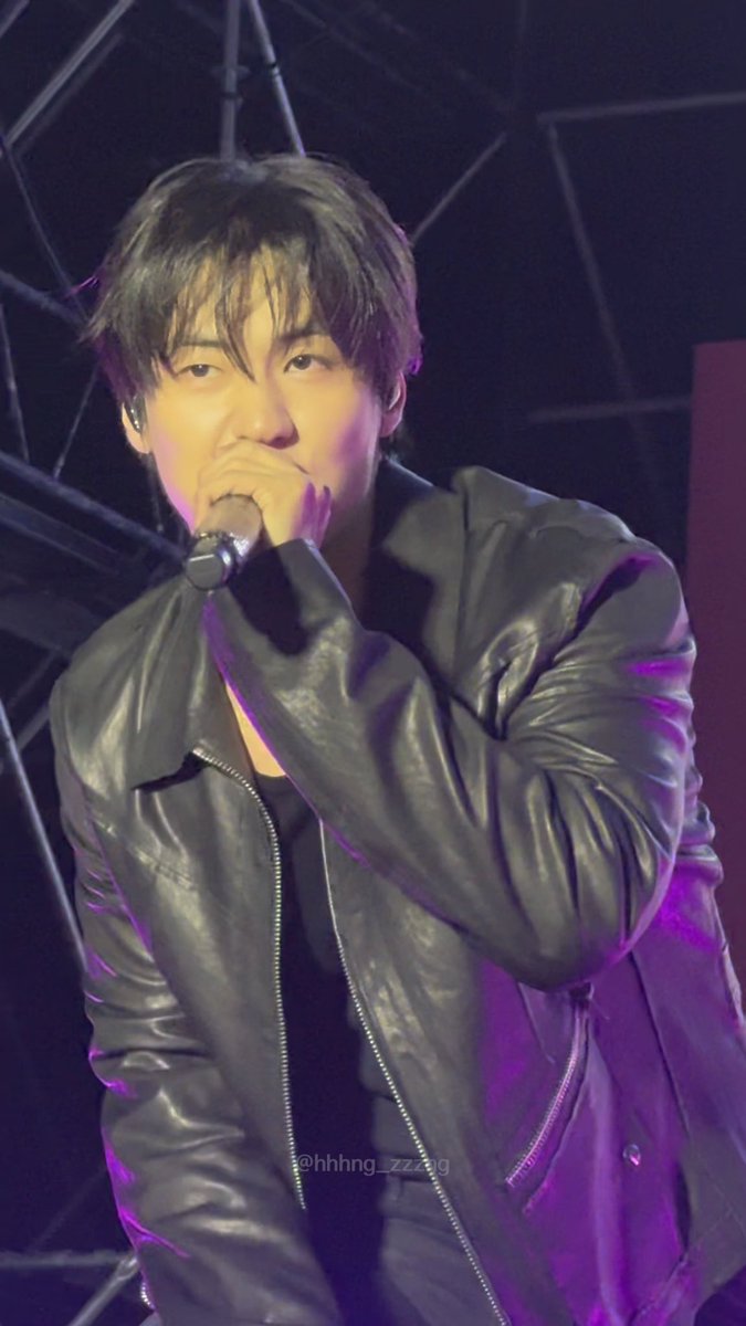 A close-up photo of a male performer in a black leather jacket singing into a microphone on stage, with a watermark '@hhhng_zzang' visible at the bottom.