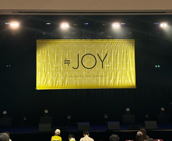 A stage banner reads '=JOY Produced by Rino Sashihara' in black text on a yellow backdrop, viewed from the audience seats.