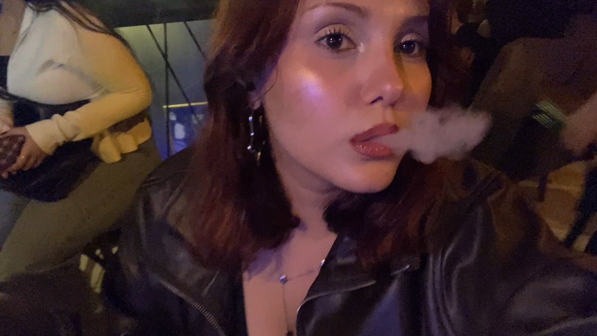 A young woman in a black leather jacket exhales vapor in a dimly lit indoor setting, likely a bar or club.