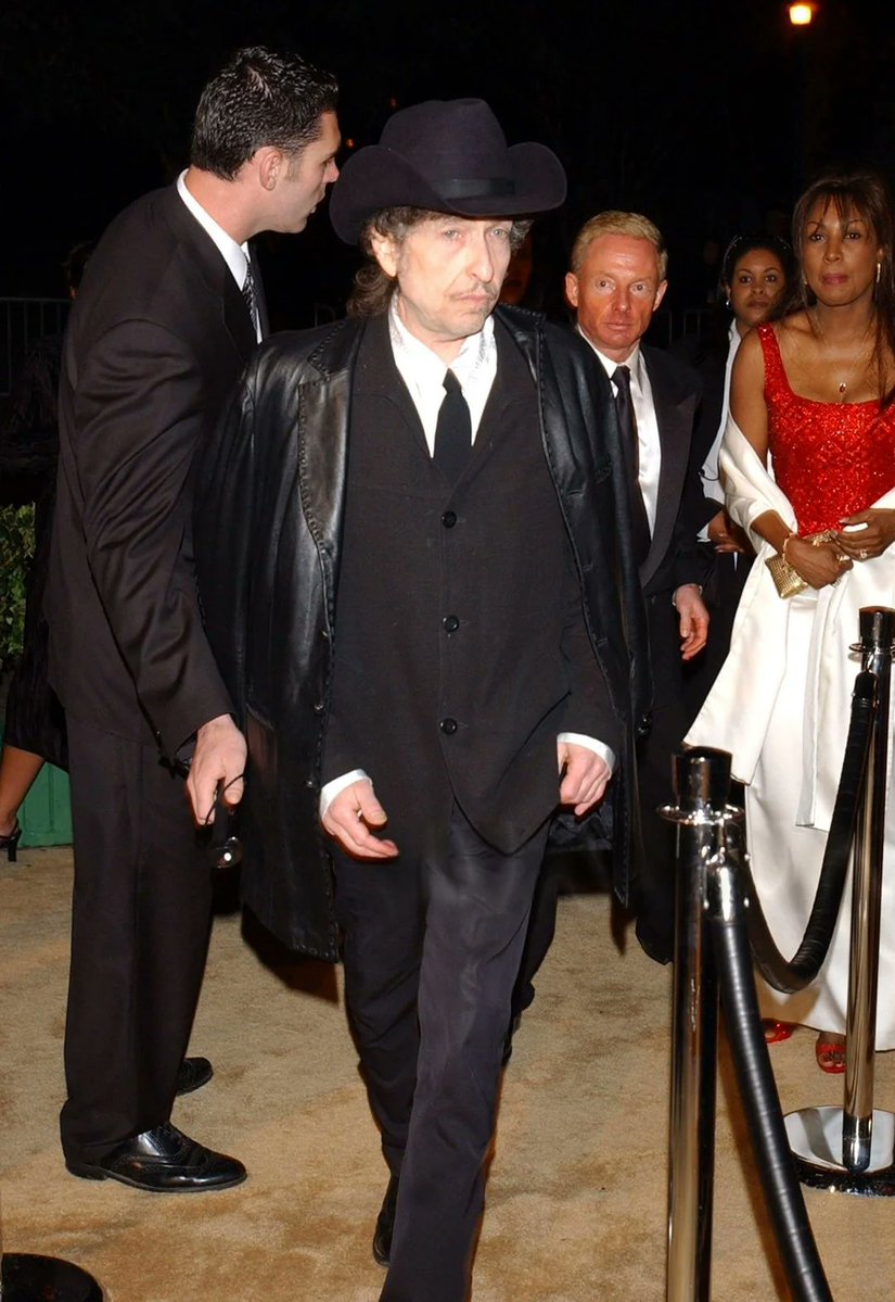 wickowolfe's tweet photo. Yıl 2002. Bob Dylan, Elton John’un geleneksel AIDS Vakfı Oscar Partisi’ne giriş yaparken kameralara yansıyor. Üzerindeki siyah deri ceketi, kovboy şapkası ve o kendine özgü, mesafeli ifadesiyle adeta bir dönem filminin içinden fırlamış gibi. Görüntü bana, Dylan’ın 1989 tarihli Oh Mercy albümünün A yüzünün kapanış şarkısı olan Man in the Long Black Coat'un atmosferini çağrıştırıyor. Şarkıdaki o gölgeli figür, karanlıkta kaybolan gizemli adam, sanki o gece o kalabalığın içinde ete kemiğe bürünmüş.  #BobDylan #ManInTheLongBlackCoat #2002 #EltonJohnParty
