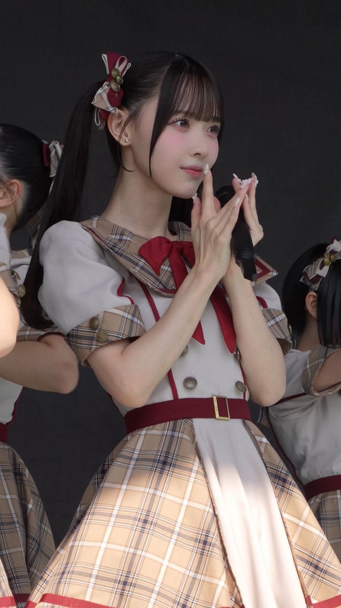 A young woman with dark hair in twin ponytails wearing a beige and red plaid idol costume holds a microphone and gestures with her hands.