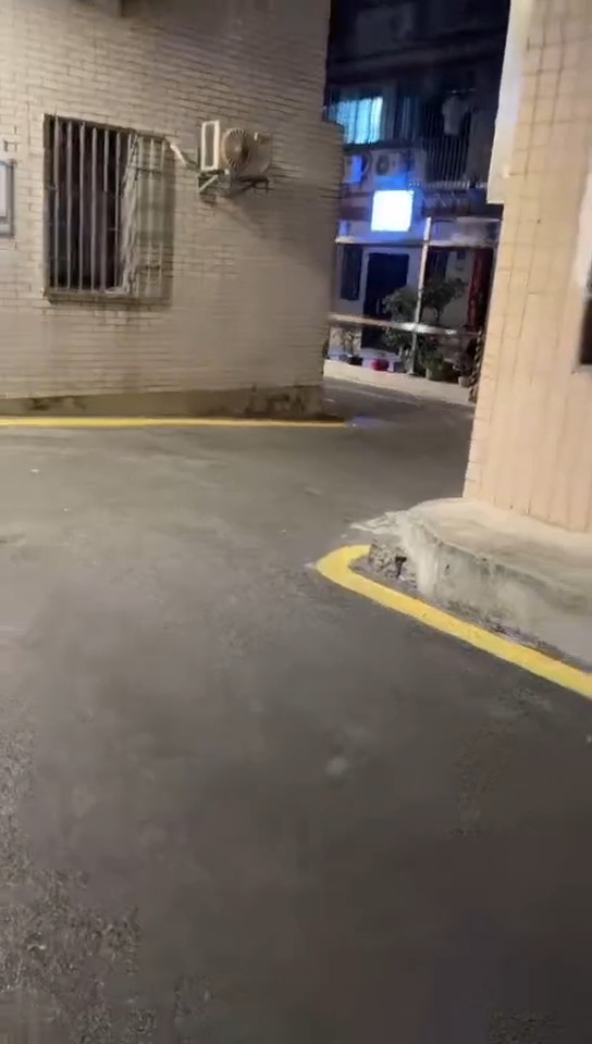 A nighttime photo of an empty narrow alleyway or street between residential buildings with barred windows, air conditioning units mounted on the tiled wall, and a yellow curb marking the edge of the pavement.