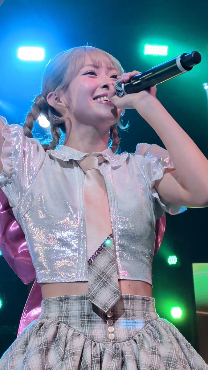 A performer with light blue hair in twin braids smiles while singing into a microphone, wearing a sparkly iridescent top with a beige tie and a plaid skirt on stage under blue and green stage lights.