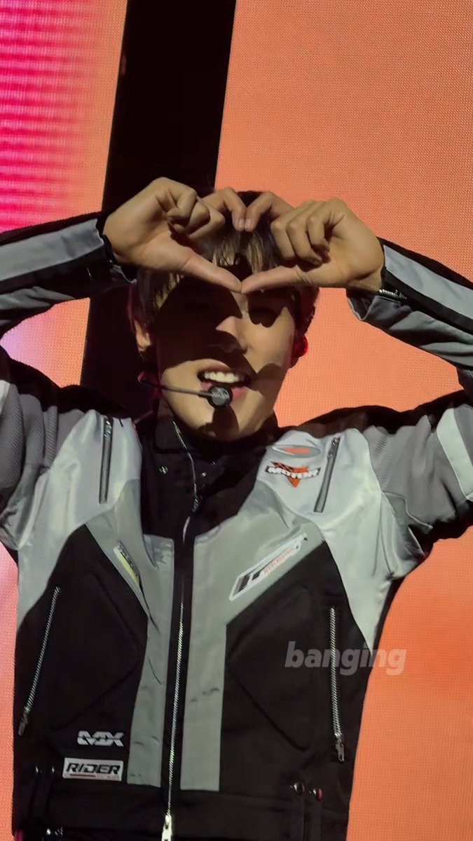 A performer on a stage with an orange background, wearing a grey/black jacket with 'MX' and 'RIDER' logos, making a heart shape with hands while wearing a headset mic; watermark 'banginimg' visible.