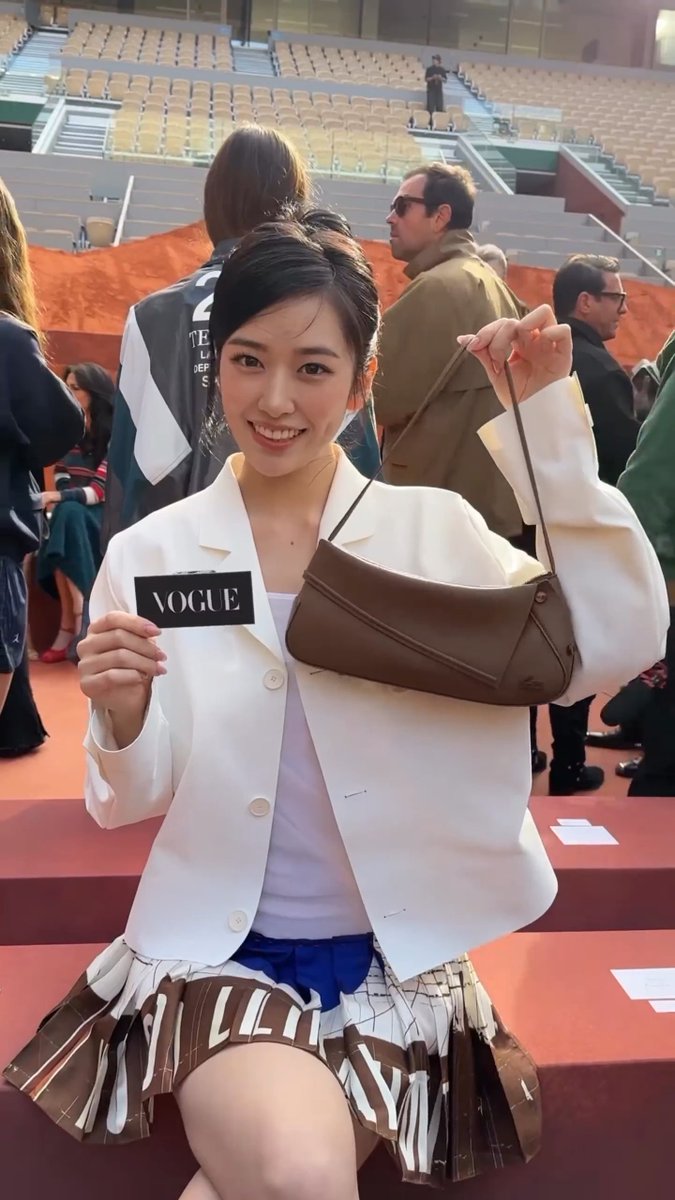 A smiling young woman at a stadium event holds a 'VOGUE' card while modeling a brown handbag, wearing a white blazer, lavender top, and pleated skirt with 'LOVE' lettering.
