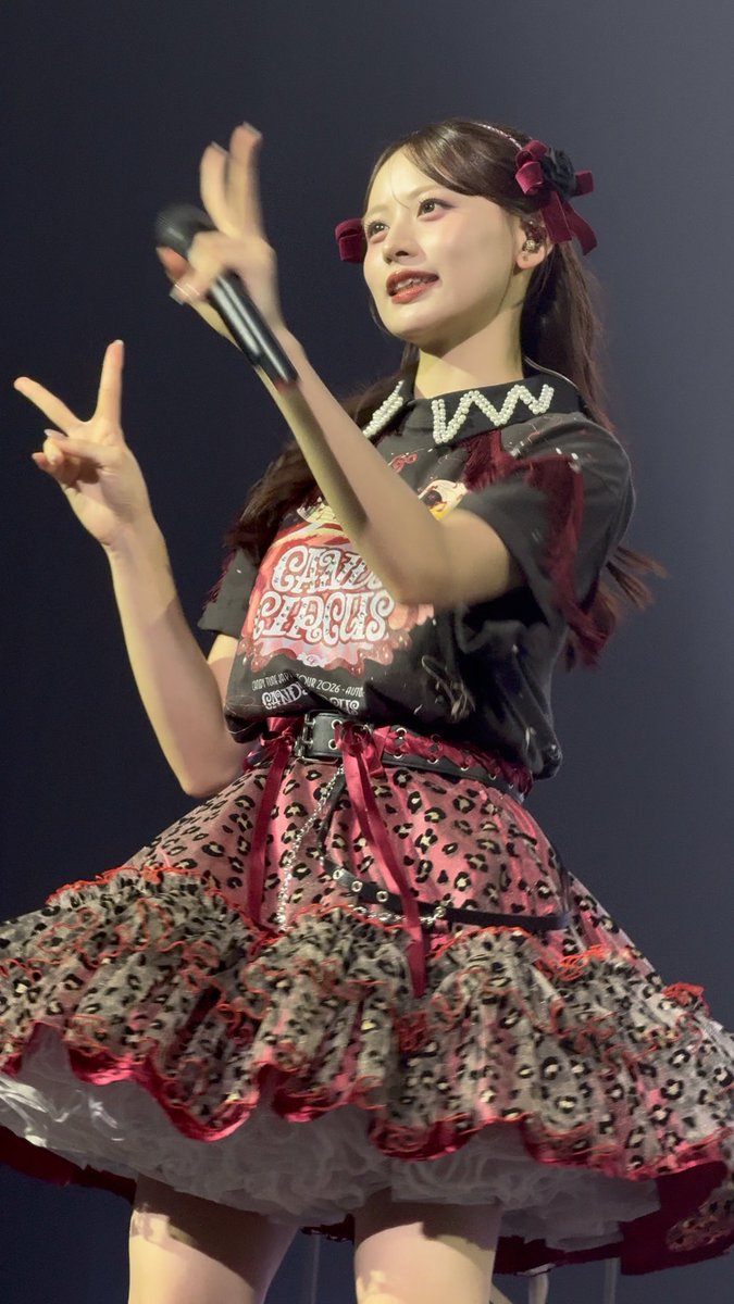 A young woman performs on stage in a red and black circus-themed outfit with 'CIRCUS' visible on her top, holding a microphone while making a peace sign.