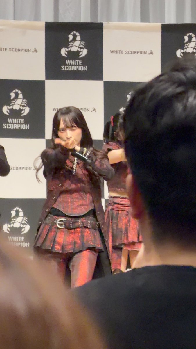 A performer sings into a microphone on stage in front of a checkered backdrop branded with the "WHITE SCORPION" logo, wearing a dark red and black ruffled outfit.