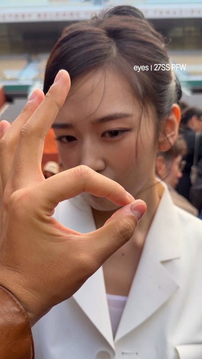 A young woman wearing a white blazer makes an 'OK' hand gesture near her face, with the text 'eyes | 27SS PFW' visible on the screen.