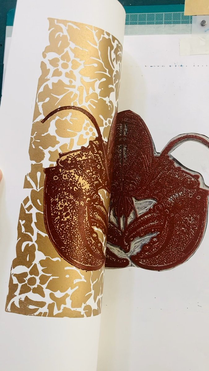 Two red rubber linocut stamps shaped like lobsters rest on a gold-and-white floral patterned cylindrical print, showcasing the carved details used to create the lobster artwork.