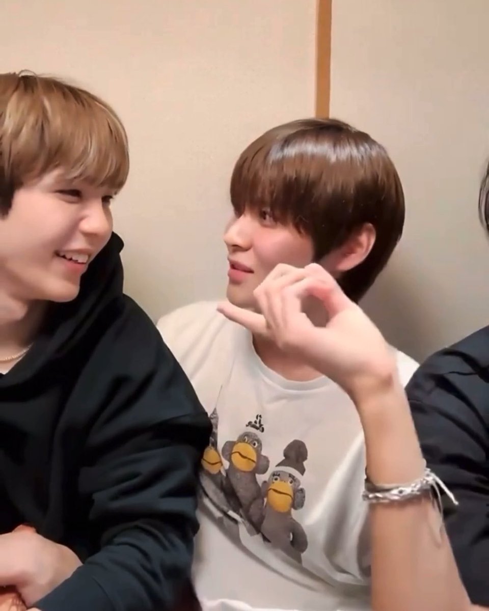 Two young men smiling at each other; the one on the right wears a white t-shirt with cartoon monkeys and makes a small gesture with his hand.