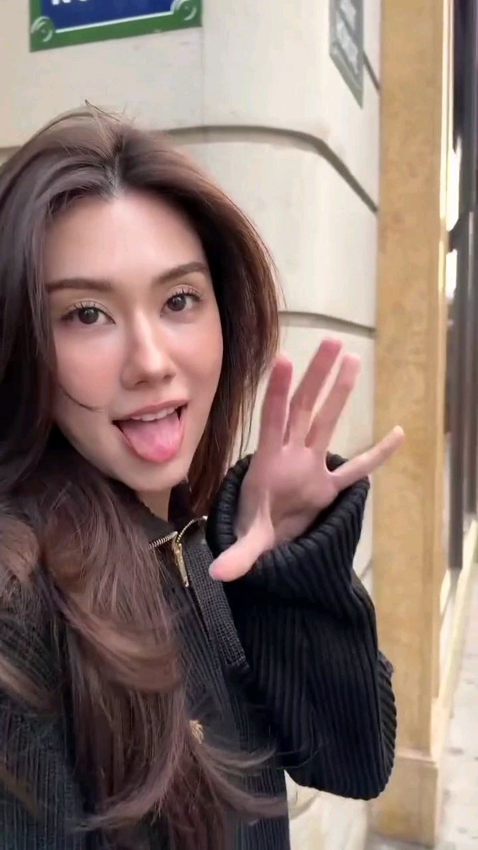 A woman with long brown hair playfully sticks her tongue out and waves at the camera while carrying a black Chanel Suede Bucket Bag with a prominent CC logo from the Cruise 2026/27 collection, photographed outside a building.
