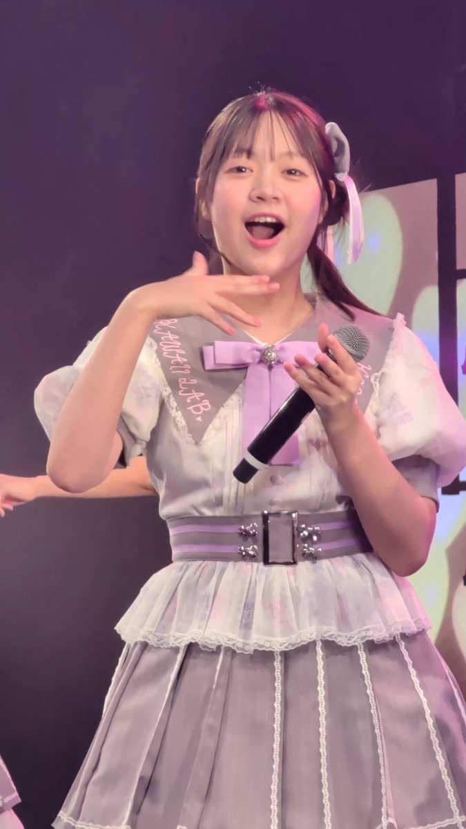 A young woman with pigtails performs on stage, wearing a grey and white dress with a purple bow, holding a microphone, with a sash reading 'B.M. 桃山台 M.B. 7月15日'.