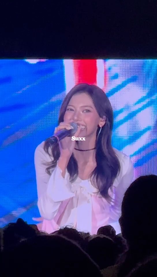 A female performer in a white blouse sings into a microphone on stage in front of a blue-lit screen, with the watermark 'Suxx' visible across the image.