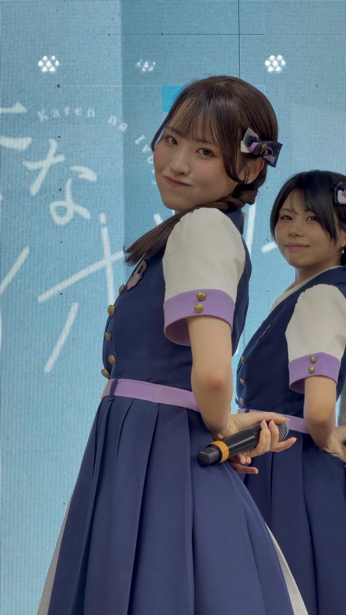 Two young women in matching navy blue and white uniforms with purple accents performing on stage, the foreground figure holding a microphone and smiling over her shoulder.