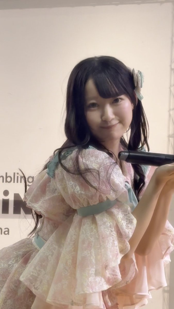 A young woman with long dark hair and a hair accessory smiles while holding a microphone, wearing a frilly, pastel-colored dress with floral patterns and light blue ribbons.