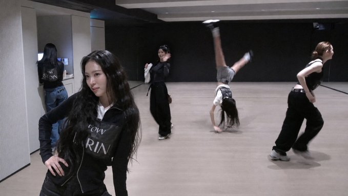 Several women practicing dance moves in a studio, with one in the foreground wearing a black jacket with 'TOM' and 'RIN' visible on it.
