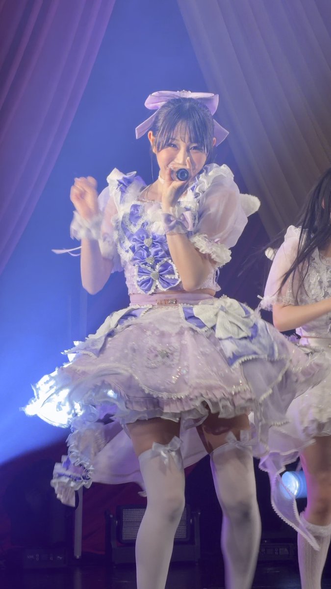 A young woman performs on stage in an elaborate lavender and white ruffled dress with a large hair bow, singing into a handheld microphone.
