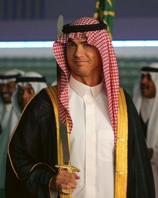 A man wearing traditional Middle Eastern attire including a red and white checkered keffiyeh and a black and gold bisht over a white thobe, holding a gold-handled sword.