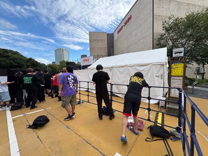 Crowd of fans queuing behind metal barriers outside a building, with one person wearing a shirt reading "ENERGY WILL WIN" and another with Japanese characters on their purple shirt.