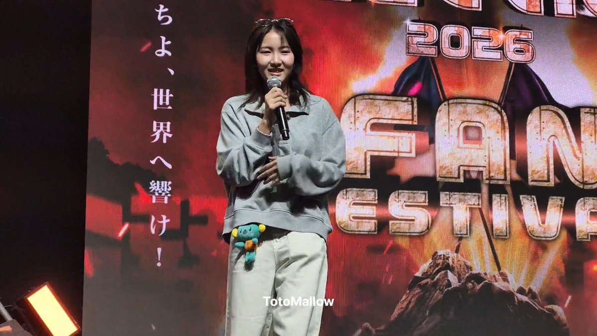 A woman in a gray sweatshirt and white pants holds a microphone while speaking at a 2026 Fan Festival event, with Japanese text and the watermark 'TotoMallow' visible.
