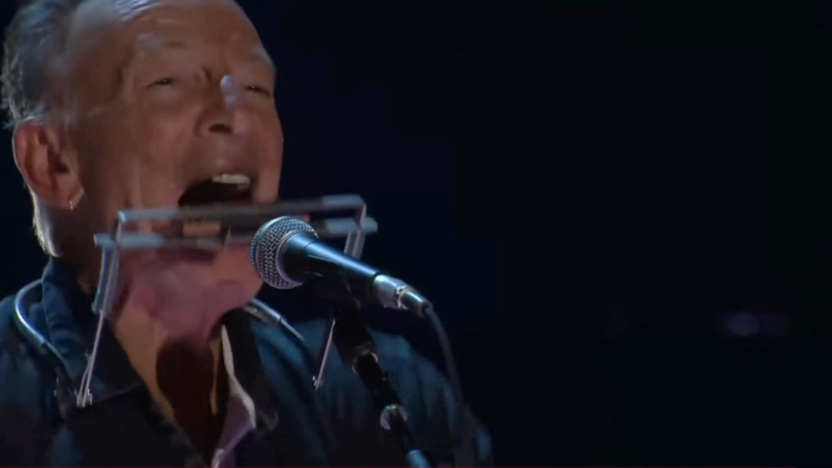 Bruce Springsteen passionately sings into a microphone while playing a harmonica on stage, captured mid-performance at what appears to be the "Power to the People" concert event organized by Tom Morello.