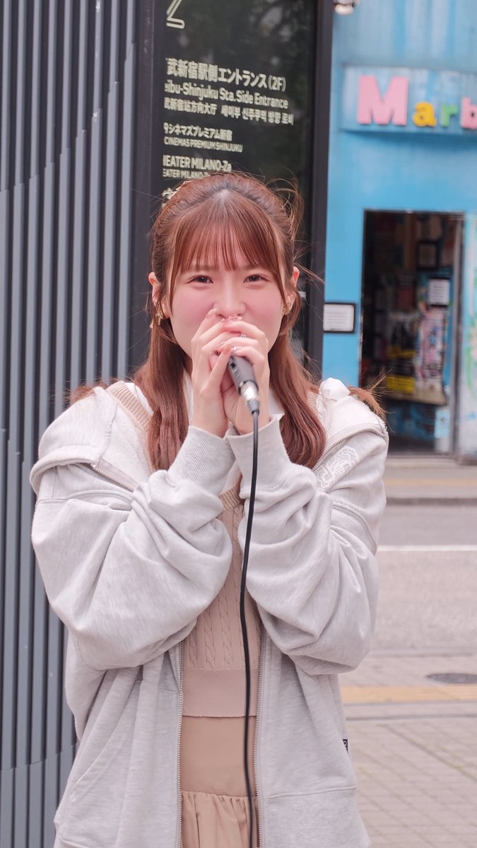 A street performer, likely a member of the Japanese idol group BABYBLOOM (ベイビーブルーム), sings into a microphone on a city street near the "Shibu-Shinjuku Sta. Side Entrance" (武蔵新宿駅東口) and "CINEMAS PREMIUM SHINJUKU" sign. Her sweet, blushing demeanor matches the group's "maybe it's mutual love" theme from the hashtag #咲けベビブル ("Bloom, Baby Bloom").