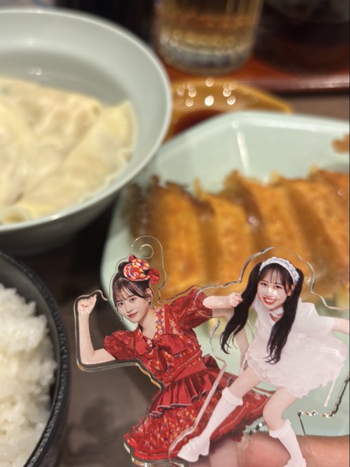 An acrylic stand featuring anime-style characters in dresses, placed in front of a meal containing rice, soup, and a breaded cutlet.