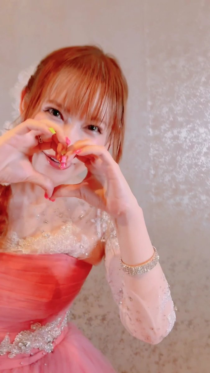 A smiling young woman with reddish-orange hair and bangs makes a heart shape with her hands while wearing a coral pink dress with sheer, embellished sleeves.