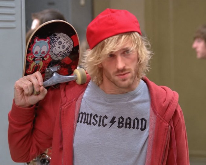 A man with shaggy blonde hair and a red backwards cap carries a skateboard over his shoulder while wearing a gray t-shirt that reads "MUSIC BAND" beneath a red hoodie.