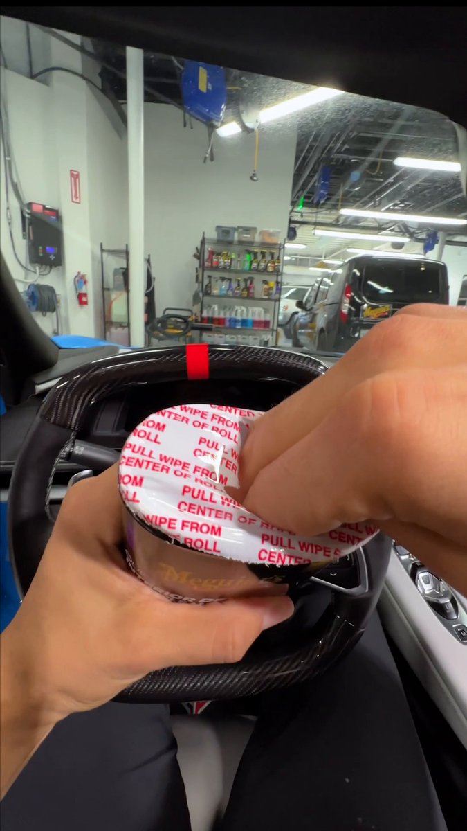 A first-person view from inside a car showing hands unwrapping a Megan brand wet wipe container, with a garage/workshop visible through the windshield.