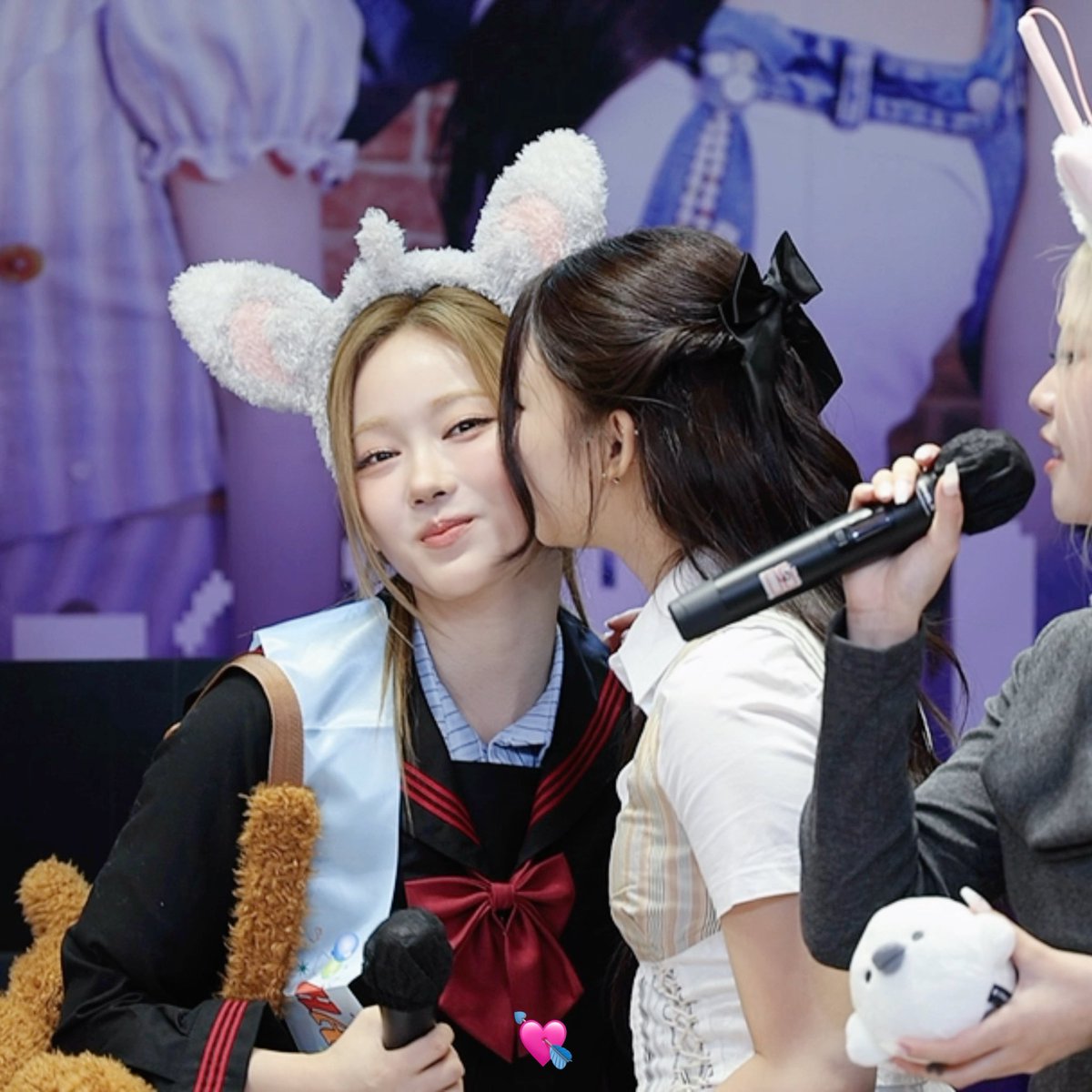 Two female idols interact on stage; one with blonde hair wears fluffy cat-ear headbands while the other leans in, and a small white plush toy is held nearby.