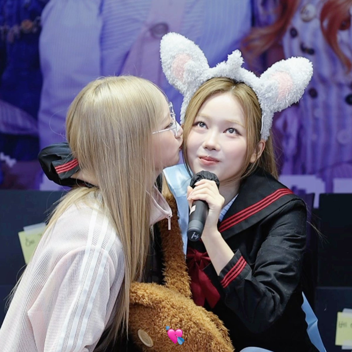 Two young women at a fan event, one wearing a sailor school uniform and the other in a dark uniform with fluffy bunny ears headband holding a microphone while being kissed on the cheek.
