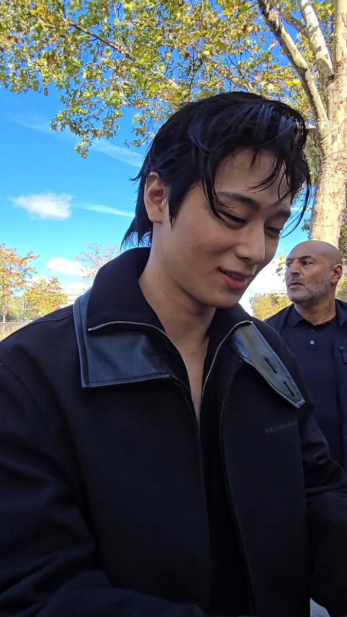 A young man with dark hair wearing a black Balenciaga jacket smiles outdoors, with a security guard in a navy polo shirt visible behind him.