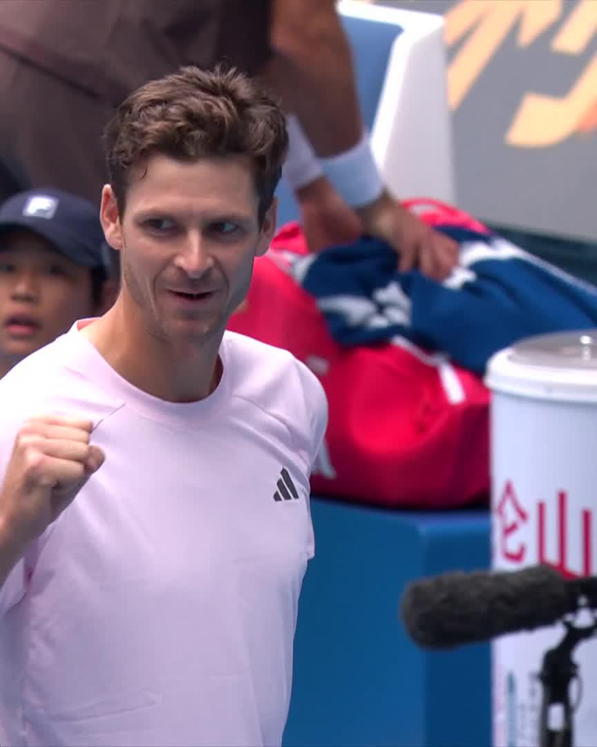 Tennis player Diego Schwartzman in a light pink Adidas shirt clenches his fist in celebration, with a Chinese sponsor logo visible on a courtside container.