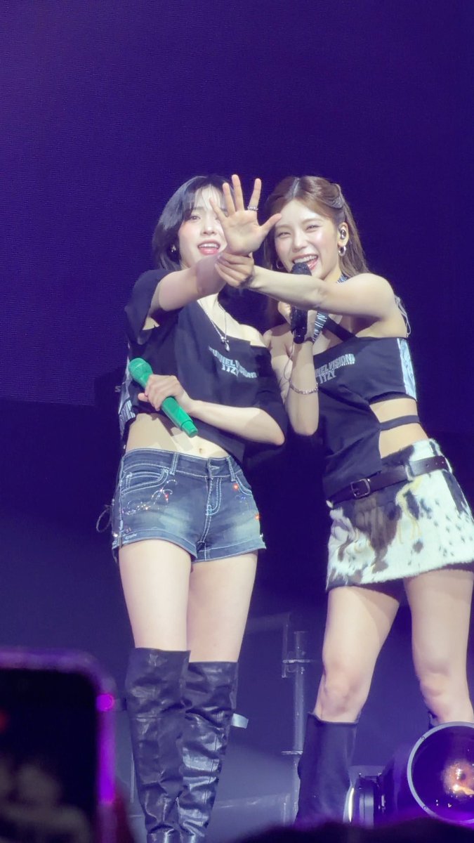 Two female K-pop artists perform on stage, both wearing black outfits; the left performer wears a denim skirt and holds a green microphone, while the right performer holds a black microphone.
