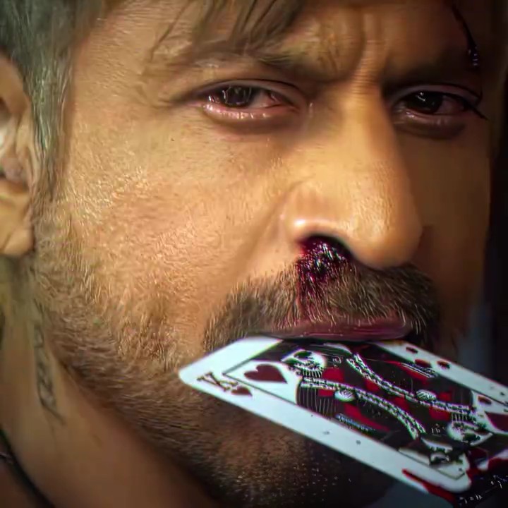 A close-up screenshot of a video game character (Johnny Silverhand from Cyberpunk 2077) holding a King of Hearts playing card in his mouth, with visible blood and a tattoo on his neck.