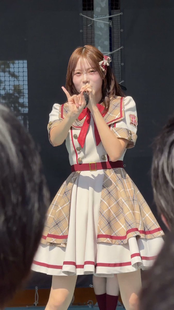A young woman performing on stage in a school uniform with a plaid skirt and red tie, making a peace sign and holding a microphone.