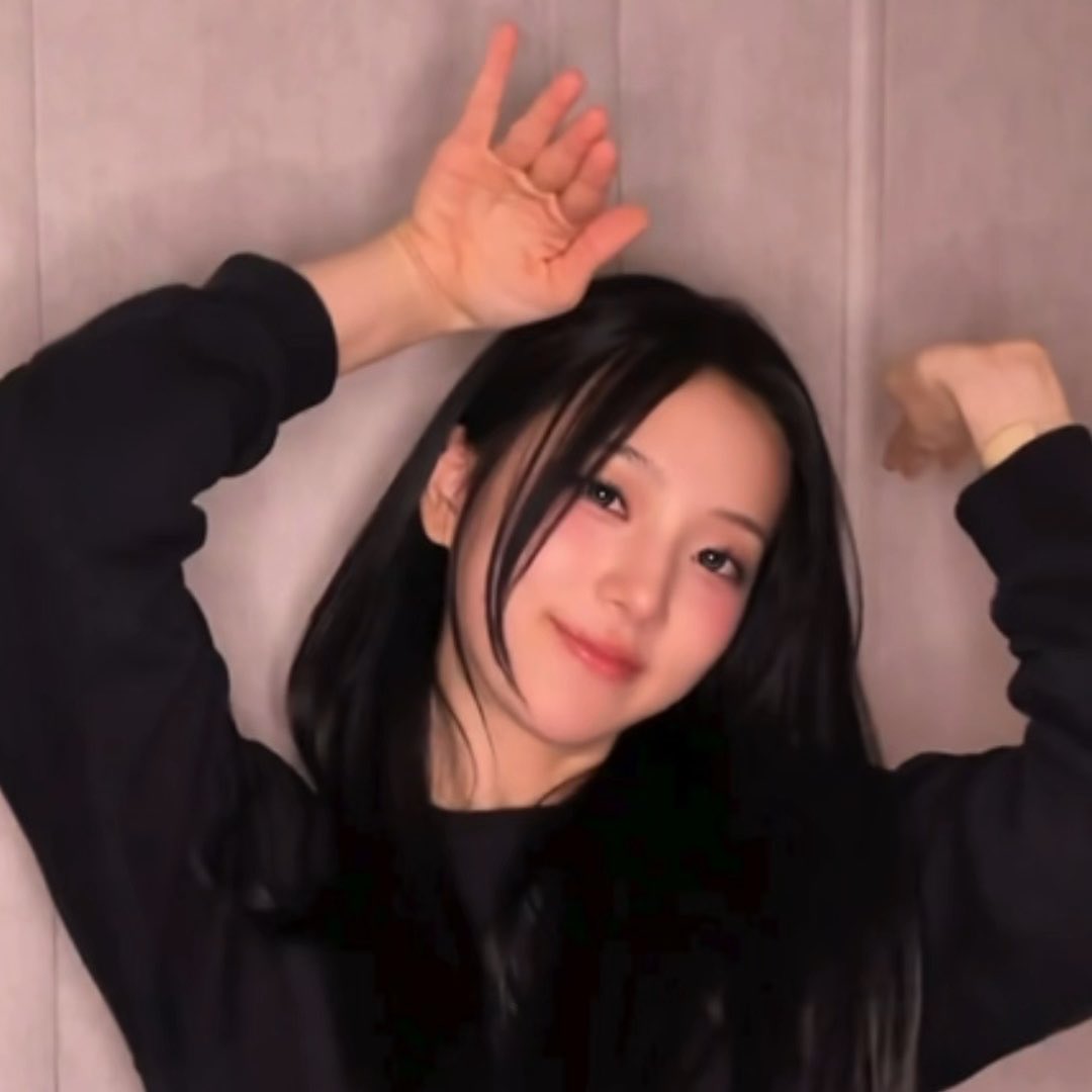 A young woman with long dark hair wearing a black top poses playfully with her arms raised, making a peace sign with one hand against a light-colored wall.