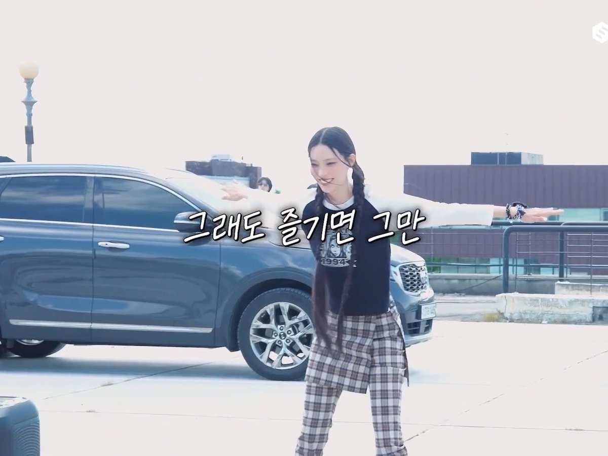 A woman with long braided hair stands in front of a car with her arms outstretched, smiling. Korean text "그래도 즐기면 그만" is overlaid on the image.
