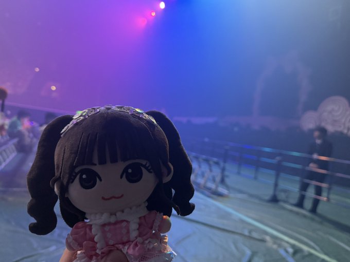A small doll with brown pigtails wearing a pink dress held up at a concert venue with blue and purple stage lighting, with an empty arena and a person walking in the background.