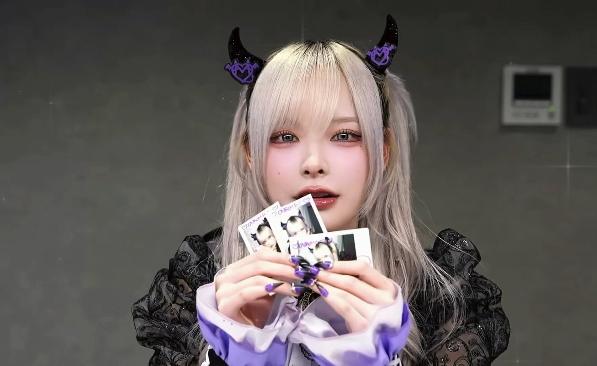 A person with silver hair and black-and-purple horn headbands holds up photos near their face, wearing a black and purple outfit with matching long nails.