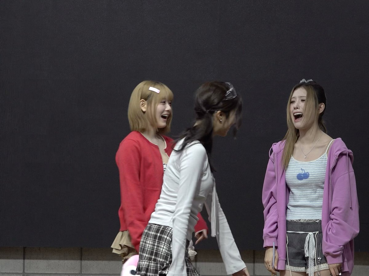 Three young women in colorful outfits (red, white, and purple) are laughing together outdoors against a dark gray background.
