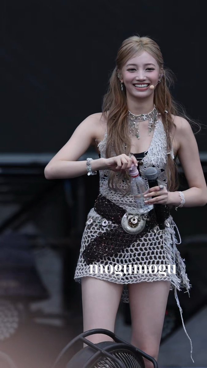 A young woman with long blonde hair, wearing a white crocheted outfit with a silver statement necklace, smiles brightly on stage with a microphone; the image is watermarked "mogumogu".