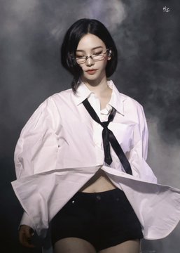 A woman with short black hair and glasses poses in a cropped white shirt with a black tie and black shorts against a dark background.